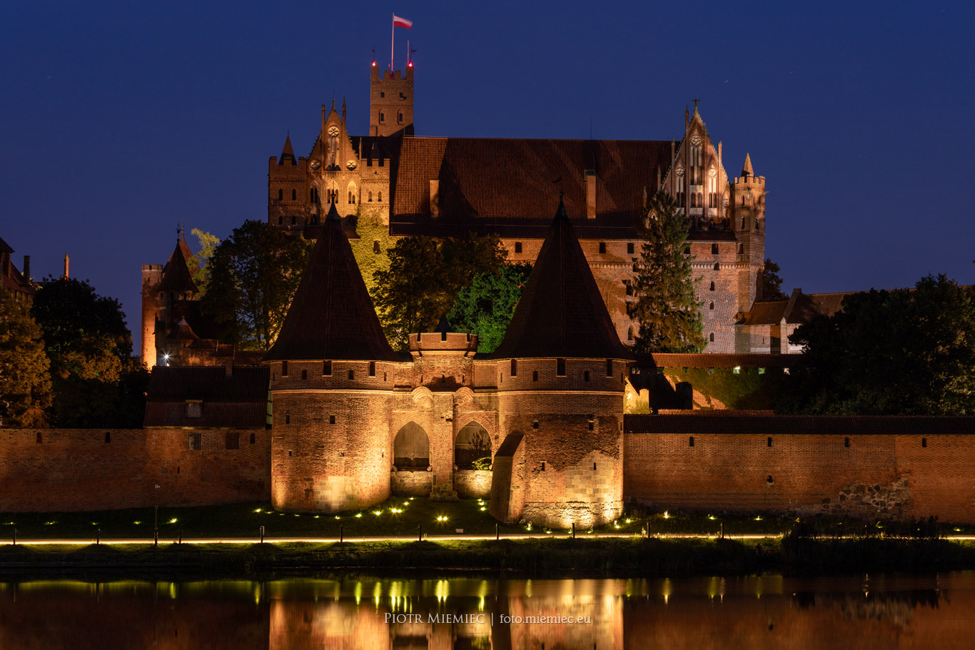 Pięknie oświetlony Zamek Malbork widziany z brzegu rzeki Nogat
