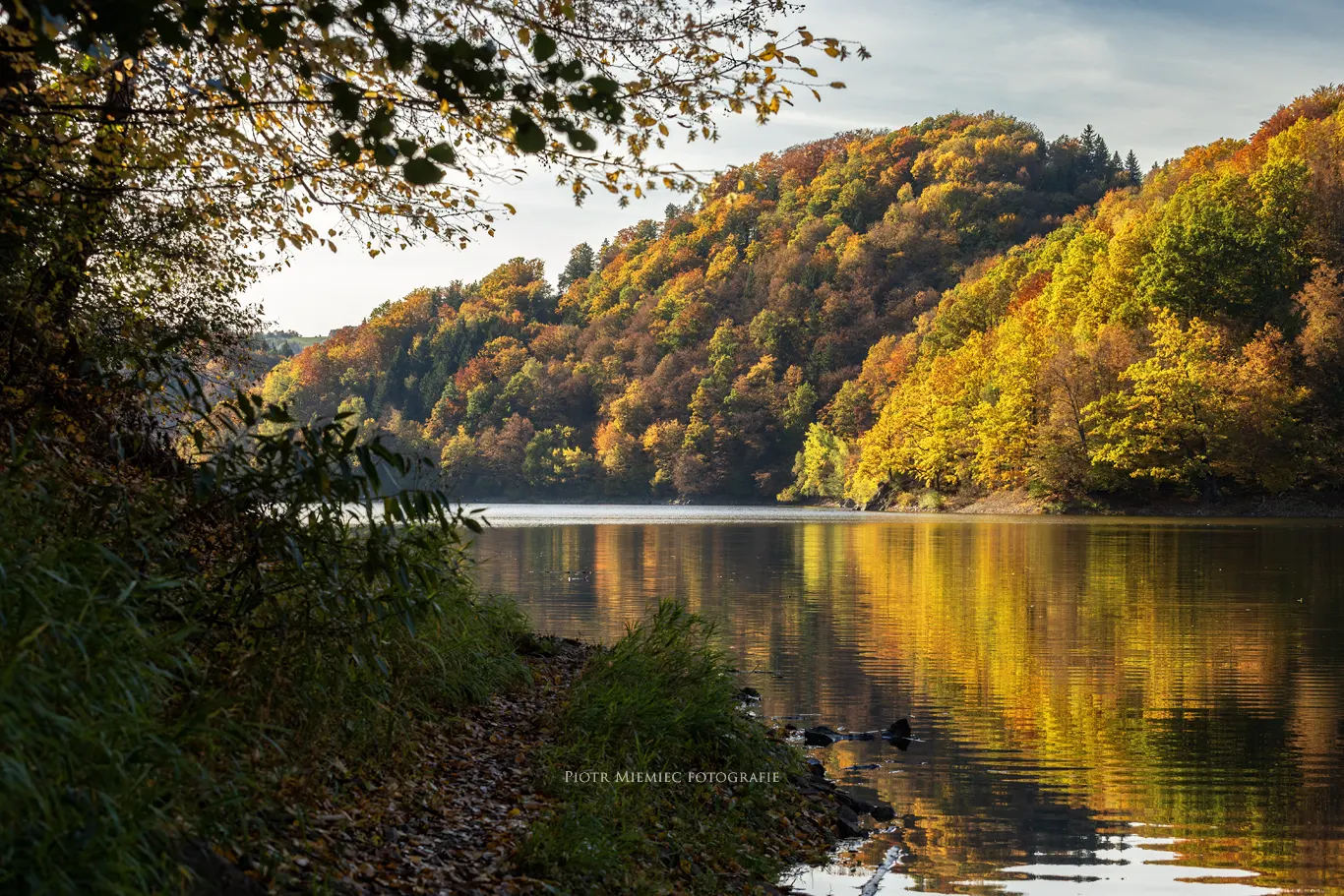 Widok na taflę jeziora z lustrzanym odbiciem jesiennego lasu.